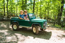 Two kids drive a small green jeep through a sunlit forest trail.