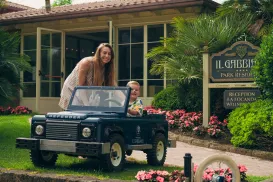 Woman and child smiling with toy car in front of a building and garden.