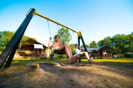 Two kids on swings in a sunny park with trees and tents in the background.