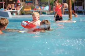 Toddler in pool with float and adults nearby, sunny day.