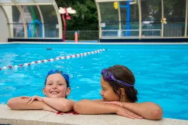 Two kids in a pool, wearing goggles, smiling by the edge.