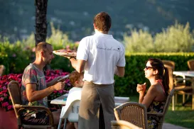 Waiter serves dishes to seated outdoor diners in sunny garden setting.