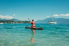 Person paddleboarding on clear blue water, distant sailboat against mountains.