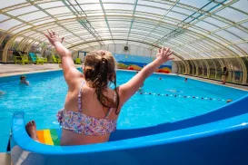 Child sliding into an indoor pool with arms raised, bright setting.
