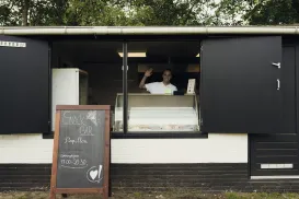 Ice cream vendor waving from a black stand with a chalkboard menu outside.