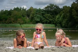 Three girls in swimsuits and sunglasses smile on a sandy beach by a river.
