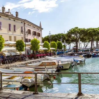 Boats docked at a quaint waterfront lined with cafes and trees under a blue sky.