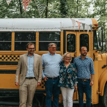 Four people smiling beside a yellow schoolbus in a natural setting.