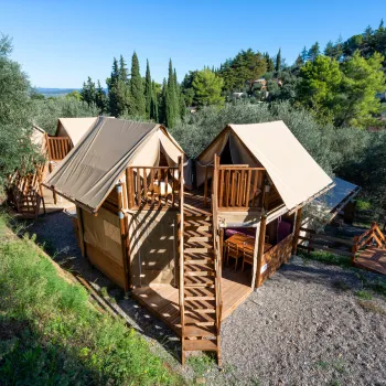 Wooden cabins with canvas roofs in a lush, hilly landscape under a clear blue sky.