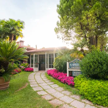 Pathway lined with pink flowers, leading to a cozy building. Trees and greenery surround it.
