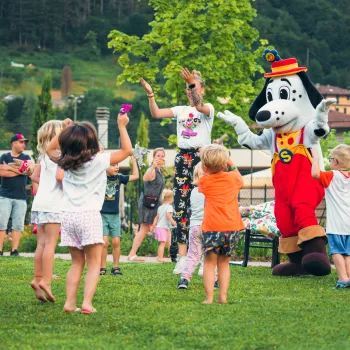 Children dancing with a costumed character in a park setting.