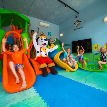 Children playing on colorful slides with a mascot in a playful, indoor setting.