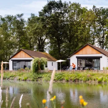 Two modern white houses by a pond, surrounded by trees and greenery.