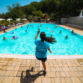 Person jumping into a crowded swimming pool on a sunny day, surrounded by trees.