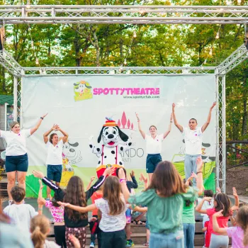 Children dancing on stage outdoors, led by adults in front of a crowd.