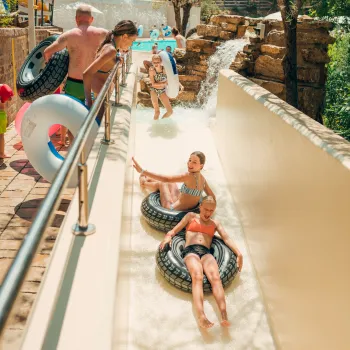 People tubing down a sunny water slide, surrounded by greenery.