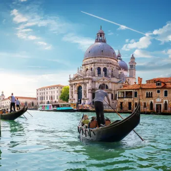 Gondolas on Venetian canal with a domed church under blue sky.