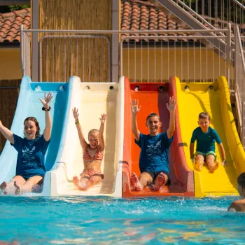 Four people joyfully sliding into a pool from colorful slides under a sunny sky.