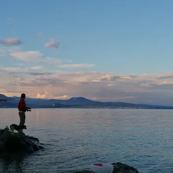 People fishing on a rock by a calm lake at sunset.