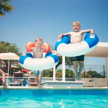 Children jumping into a pool with floatation rings, under a clear blue sky.