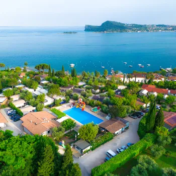 Aerial view of a lakeside resort at lake garda with pool, trees, and distant island.