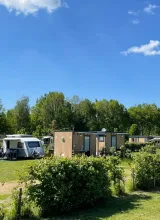 Grass field with camper vans, wooden cabins, and trees under a blue sky.