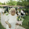 Elderly woman in white dress holding flowers, smiling outdoors at a garden event.