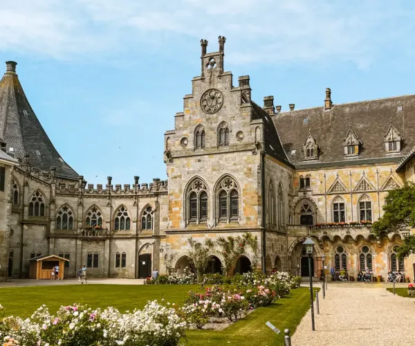 Historic stone building with clock tower, surrounded by gardens, under a blue sky.