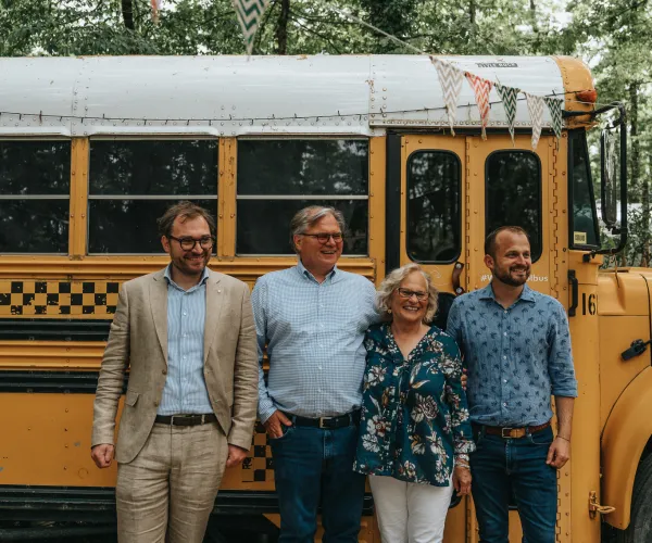 Four people smiling beside a yellow schoolbus in a natural setting.
