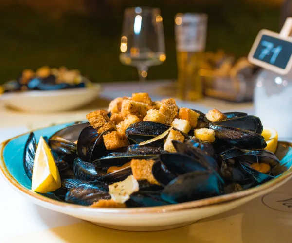 Bowl of mussels with croutons and lemon slices on a table with wine and a beer glass.