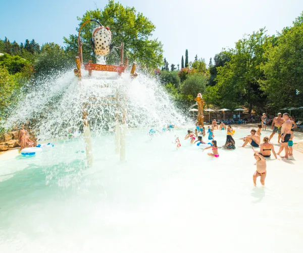 Water play area with a large splash bucket tipping over kids, surrounded by trees.