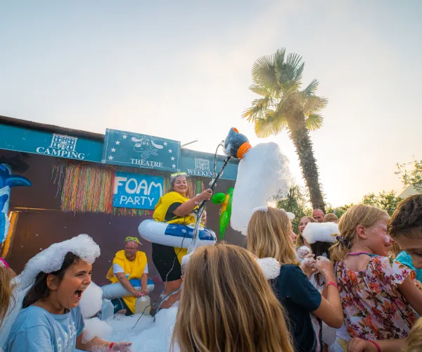 Foam party with smiling children, palm tree in background, colorful and lively atmosphere.