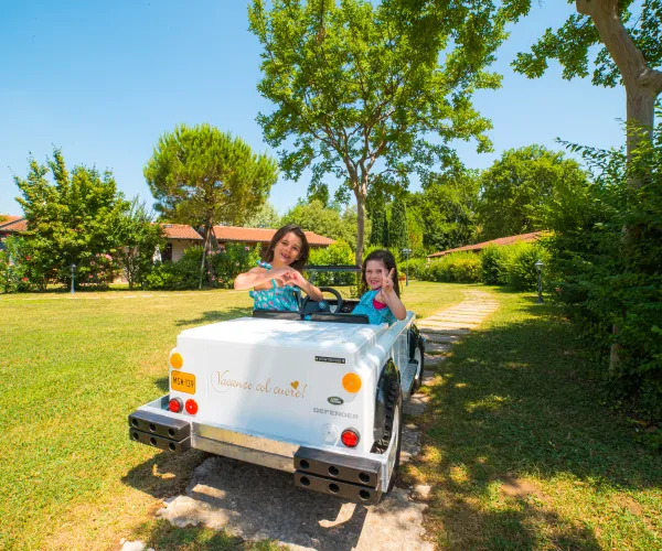 Two people in a toy car on a sunny day, surrounded by trees.