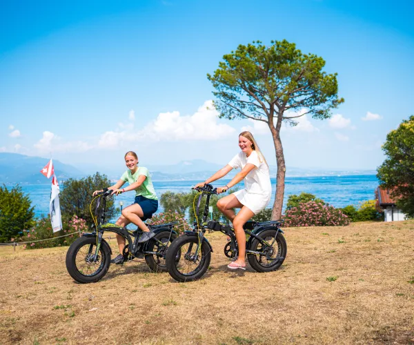 Two people ride e-bikes on a grassy hill by a lake, under a clear blue sky.