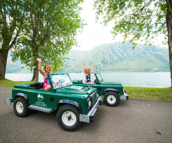 Children driving toy cars on a lakeside road, smiling and waving.
