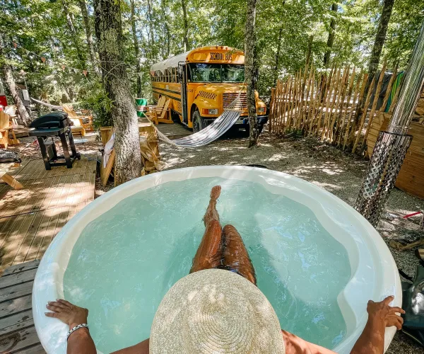 Person relaxing in a hot tub, wearing a straw hat, with trees and yellow bus in the background.
