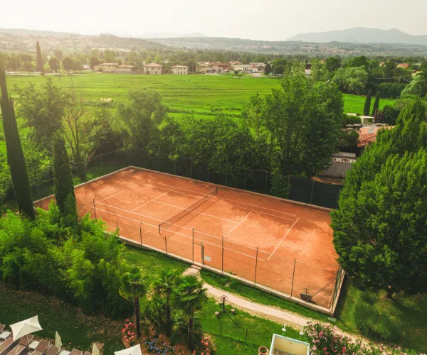 Clay tennis court surrounded by lush greenery, with distant hills and houses.