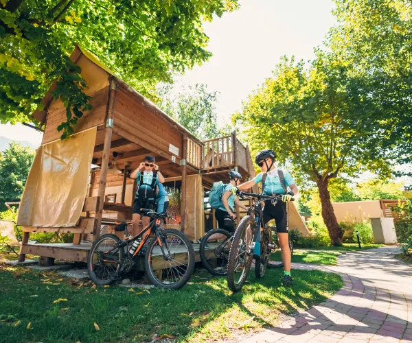 Cyclists resting by a wooden cabin, surrounded by trees on a sunny day.