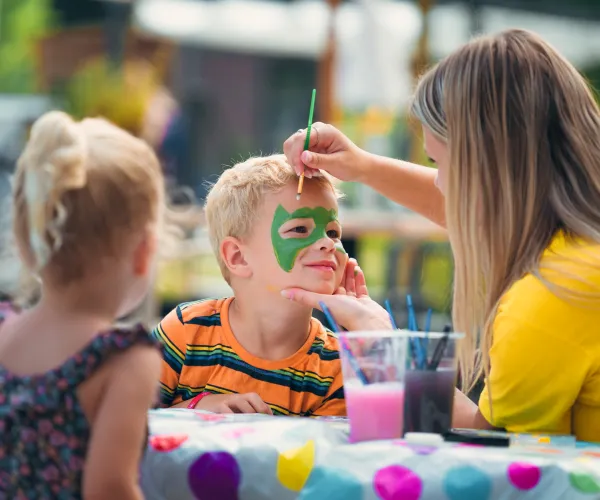 Child getting face painted green by an adult at a table, another child watching.