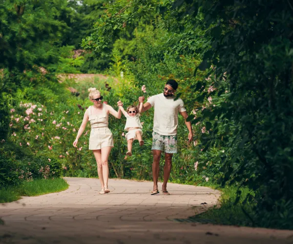 Family walking on a path in a lush, green park, lifting a child between them.