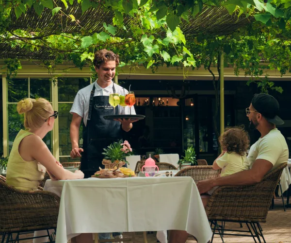 Waiter serves drinks to a family at an outdoor restaurant table.