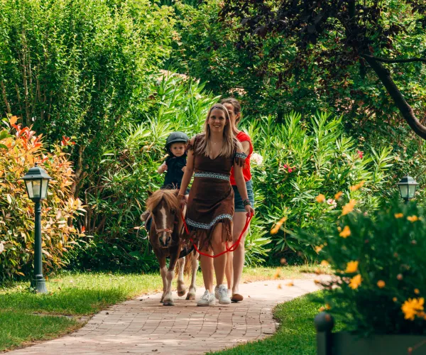 Child riding a pony with two adults guiding in a lush garden path.