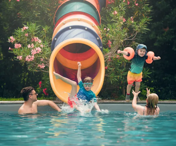 Children sliding into pool, adults catching them, colorful slide in background.