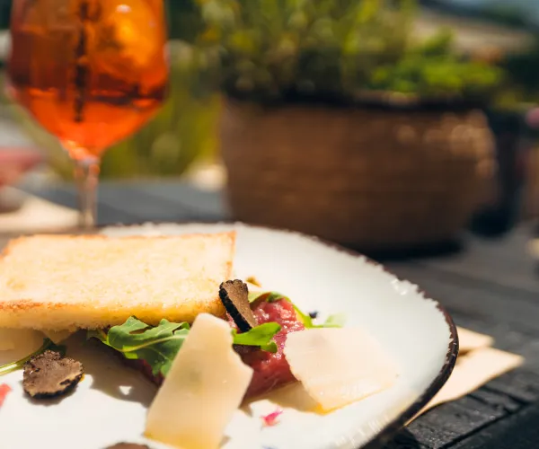 Toasted bread with arugula, shaved parmesan, truffles on a white plate, glass of iced tea nearby.