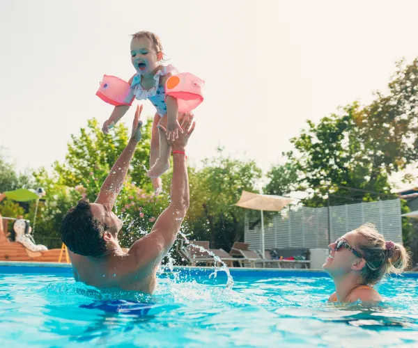 Father lifts child in pool with mother nearby, sunny day.