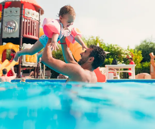 Father lifts child in pool, smiles, colorful playground in background.