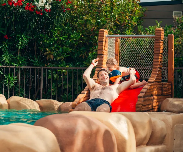 Man and child sliding down a water slide, surrounded by lush greenery.