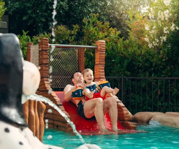 Father and child on water slide, smiling in a pool with greenery around.