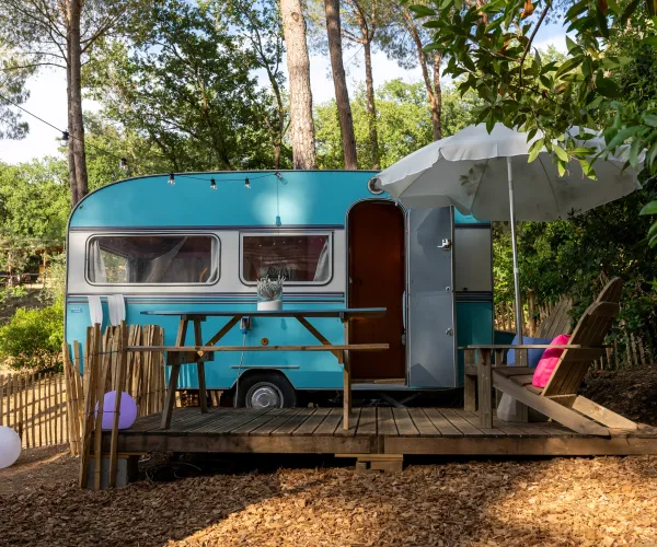Blue vintage camper in a wooded setting with a deck, parasol, and chairs.