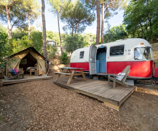 Retro camper and tent in a wooded campsite with picnic tables.